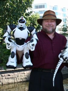  A photograph of Mark Tilden wearing a wide brimmed hat, holding a white Robosapien, an approximately one foot high humanoid robot, in one hand. He is standing outside with an office building in the background. 