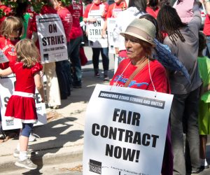  A photograph of a teacher protesting in a crowd, with a large, square sign around her neck reading “Fair Contract Now!”