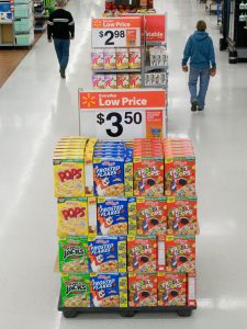 A sale display in a Wal-Mart aisle. The display shows four different types of cereal stacked beside each other, with a price sign that reads “$3.50” sitting on top. In the background are other sales displays and several customers walking in the aisle.