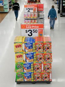  A sale display in a Wal-Mart aisle. The display shows four different types of cereal stacked beside each other, with a price sign that reads “$3.50” sitting on top. In the background are other sales displays and several customers walking in the aisle. 