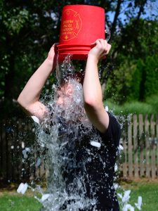 A photograph of a person standing outside, holding a red bucket upside down above their head. Out of the bucket, water and ice is falling on the person, obscuring their face and upper torso.