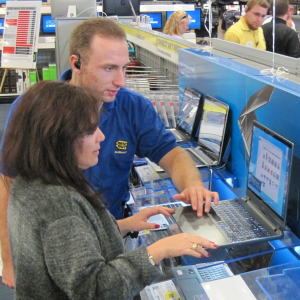 A photograph of a Best Buy employee showing a customer a computer. The employee and customer are standing in front of a computer on a display, with the employee showing the customer a page on the laptop. In the background is the rest of the Best Buy store, with other customers and employees.