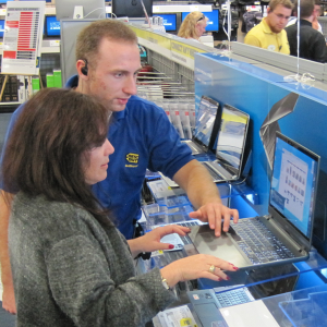  A photograph of a Best Buy employee showing a customer a computer. The employee and customer are standing in front of a computer on a display, with the employee showing the customer a page on the laptop. In the background is the rest of the Best Buy store, with other customers and employees.