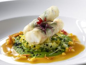 A photograph of what appears to be two spring rolls, sitting on top of a layer of rice with green and orange sauce. Red flowers accent the spring rolls. The food sits on top of a white plate, on a white background.