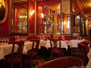  A photograph of the inside of Le Procope. Four tables with white tablecloths are in the foreground, with wooden chairs around them. Behind them on the wall are mirrors and a display of glasses. 