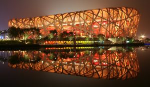  A photograph of the Beijing National Stadium at night, taken from across water. The National Stadium is lit up by yellow lights from within. A reflection of the stadium appears on the water. 