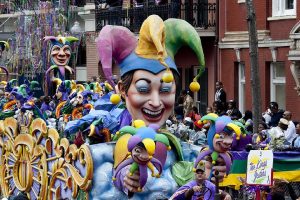  A photograph of a parade during Mardi Gras in New Orleans. The photograph is centered on a parade float shaped like a jester holding two smaller jesters. All three jesters have purple, yellow, and green hats. More identical jesters are in the background, along with a crowd of people and brick buildings. 