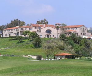 A photograph of a very large, two-story Spanish style mansion on top of a hill, with trees in the background and in front of the mansion. Another smaller house with a red roof sits at the bottom of the hill.