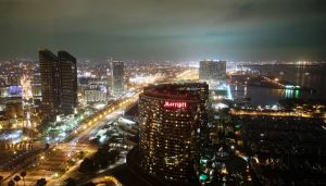  A photograph of the San Diego cityscape at night, lit up with lights. The San Diego Marriott in the foreground, a large, arch shaped skyscraper. Water is to the right of the Marriott, with lighted docks extending into the water. A highway is to the left, lined with streetlights. Other buildings are in the background, including two other skyscrapers to the left and two in the distance.