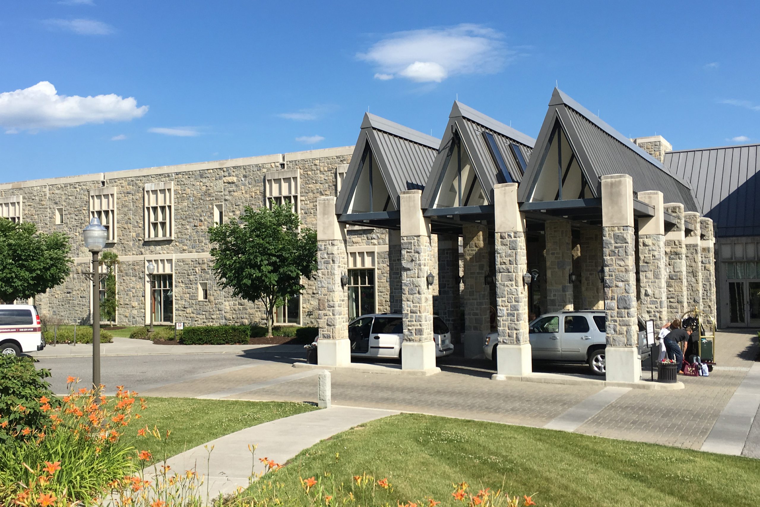  A photograph of the front entrance of The Inn at Virginia Tech, shown from the front with a path leading up to the entrance. The building is rectangular and two stories high, with windows on each level. The front entrance features four stone columns, with three triangles forming the roof. A white car sits under the roof, where people are unloading bags.