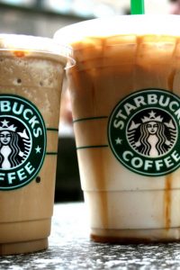 A photograph of two Starbucks drinks beside each other in clear cups on a table, with the Starbucks icon visible on both cups. The cup on the left is a smaller size and filled with light brown frozen coffee. The right cup has coffee in the top and cream in the bottom, with a gradient in between.