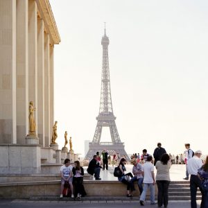 Left side of the picture shows the right side of a building in the foreground with many people. In the background is the Eiffel Tower in Paris, France.