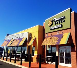 A picture of a Banera Bread restaurant exterior. Colorful awnings and paint on the building. A sign that indicates that this restaurant has a drive thru.