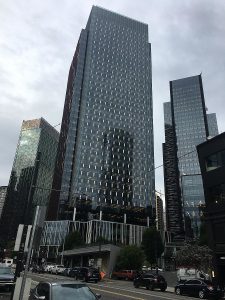 Three dark gray reflective skyscrapers sit on a busy road. The picture focuses on one skyscraper up close with the other 2 in the background. The sky is light gray and cloudy. Fifteen vehicles sit out front parked on the street.