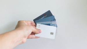Picture of a hand holding 4 credit cards fanned out in their hand. The cards are Capital One, Discover, American Express, and Apple. The background is white.