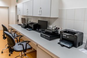 An office space with a desk that holds two computer monitors and two printers, both are black.