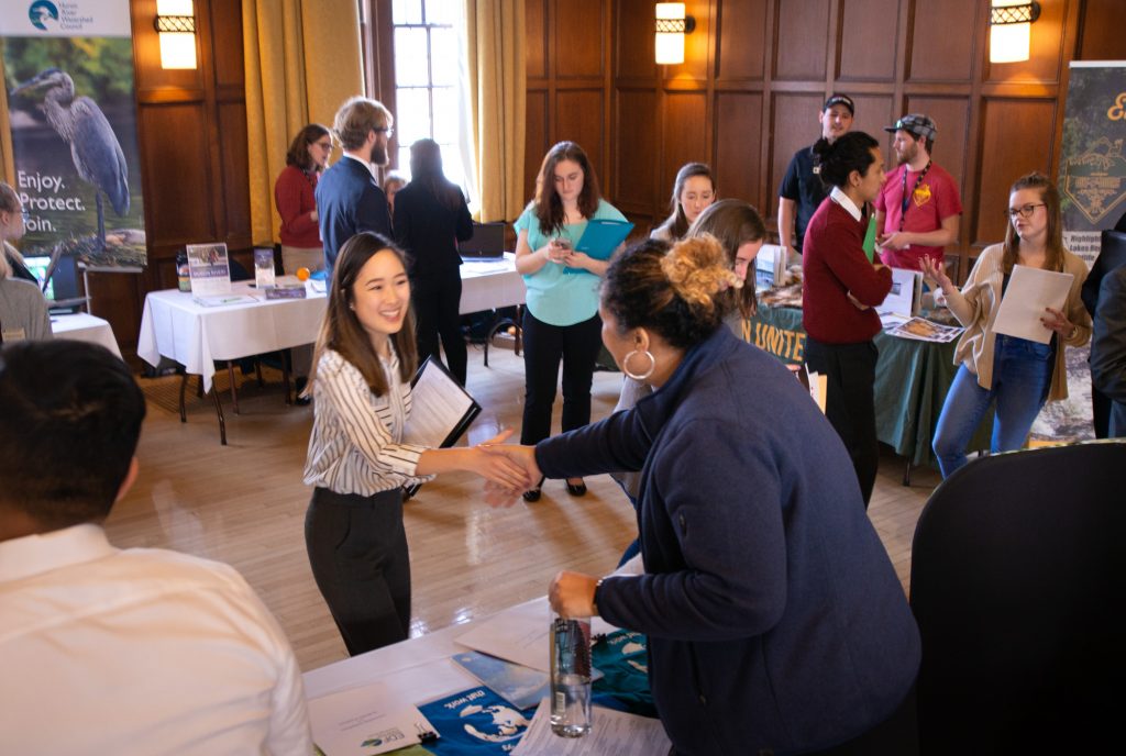 A typical college career fair with tables set out in a large conference room. Two women are skaking hands across one of the tables.