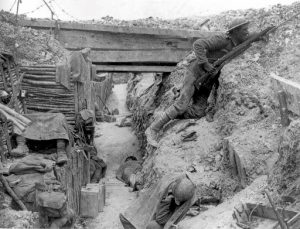 Black and white photo inside of a trench in 1916 during the Battle of Somme. At least 4 men lay in the trench while one is holding a rifle still fighting.
