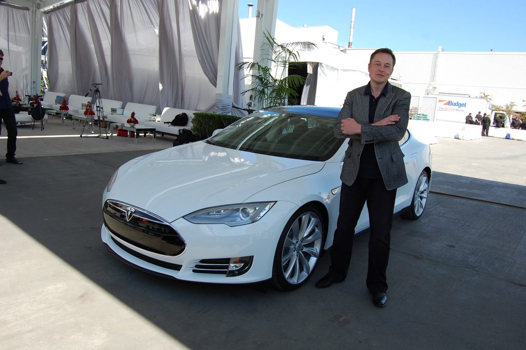 Elon Musk standing next to a Tesla sportscar outside the Tesla factory.