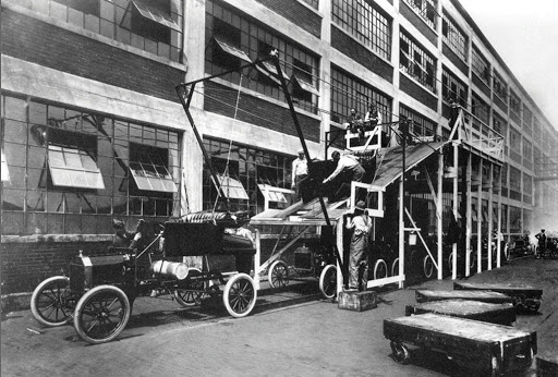 Photograph from 1913 of For'd assembly line. A car is being assembled outside of a factory.