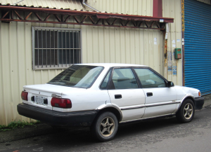 Old white Geo brand car parked outside of a building.