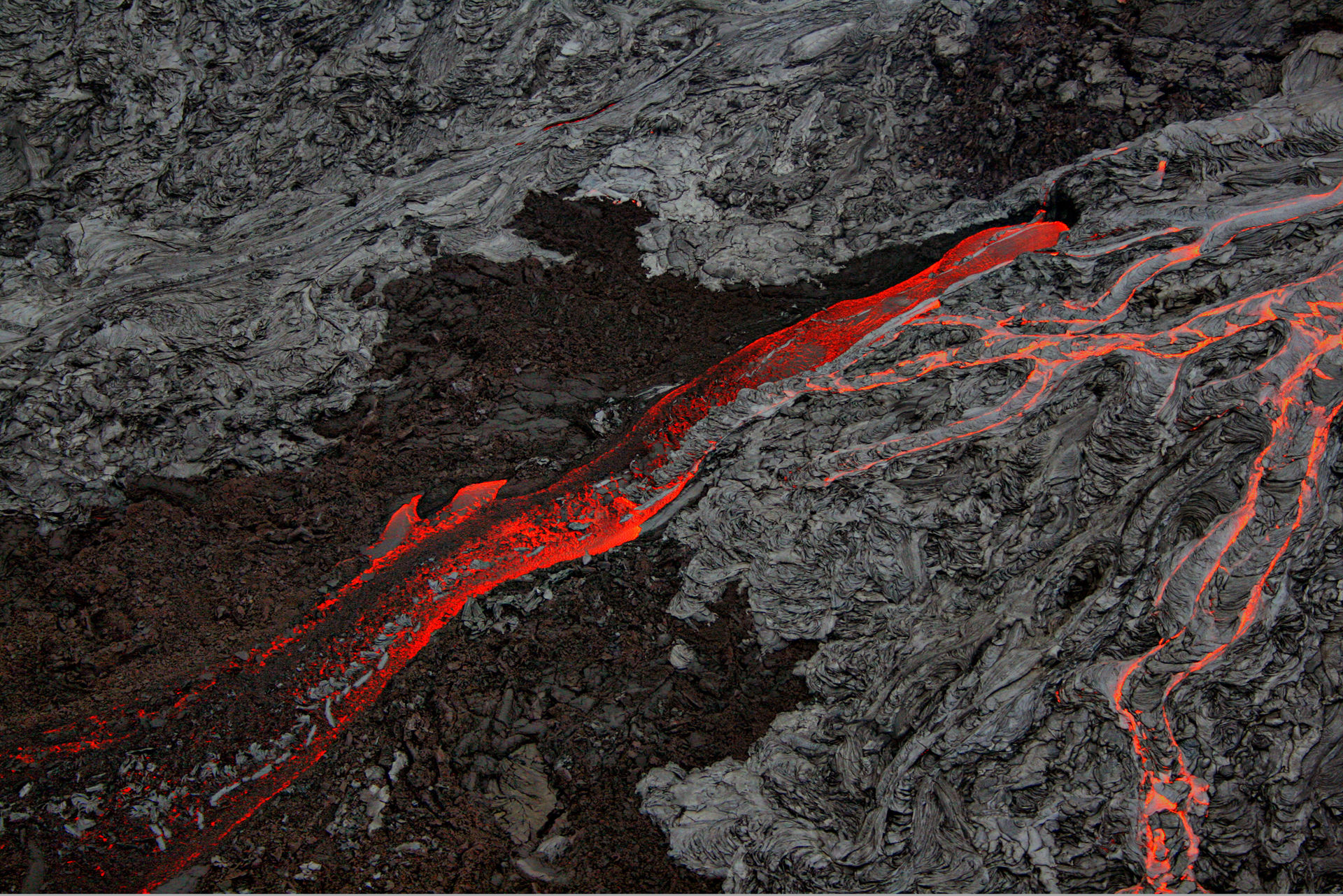 Stream of orange molten lava traveling down gently sloping hill of cooled black and gray volcanic rock.