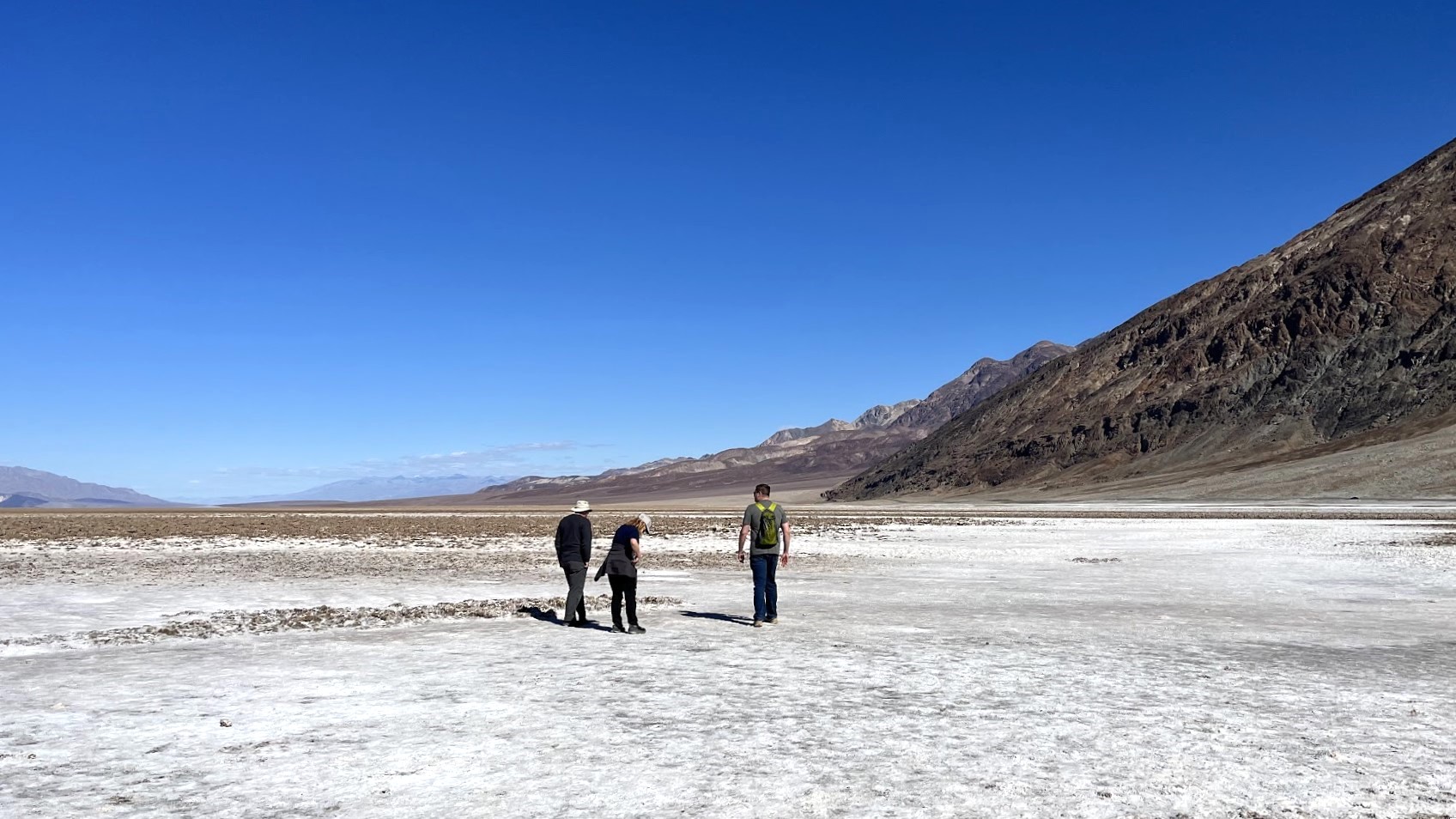 Three people standing on a flat white expanse with a steep hill rising to the right side of the photo.