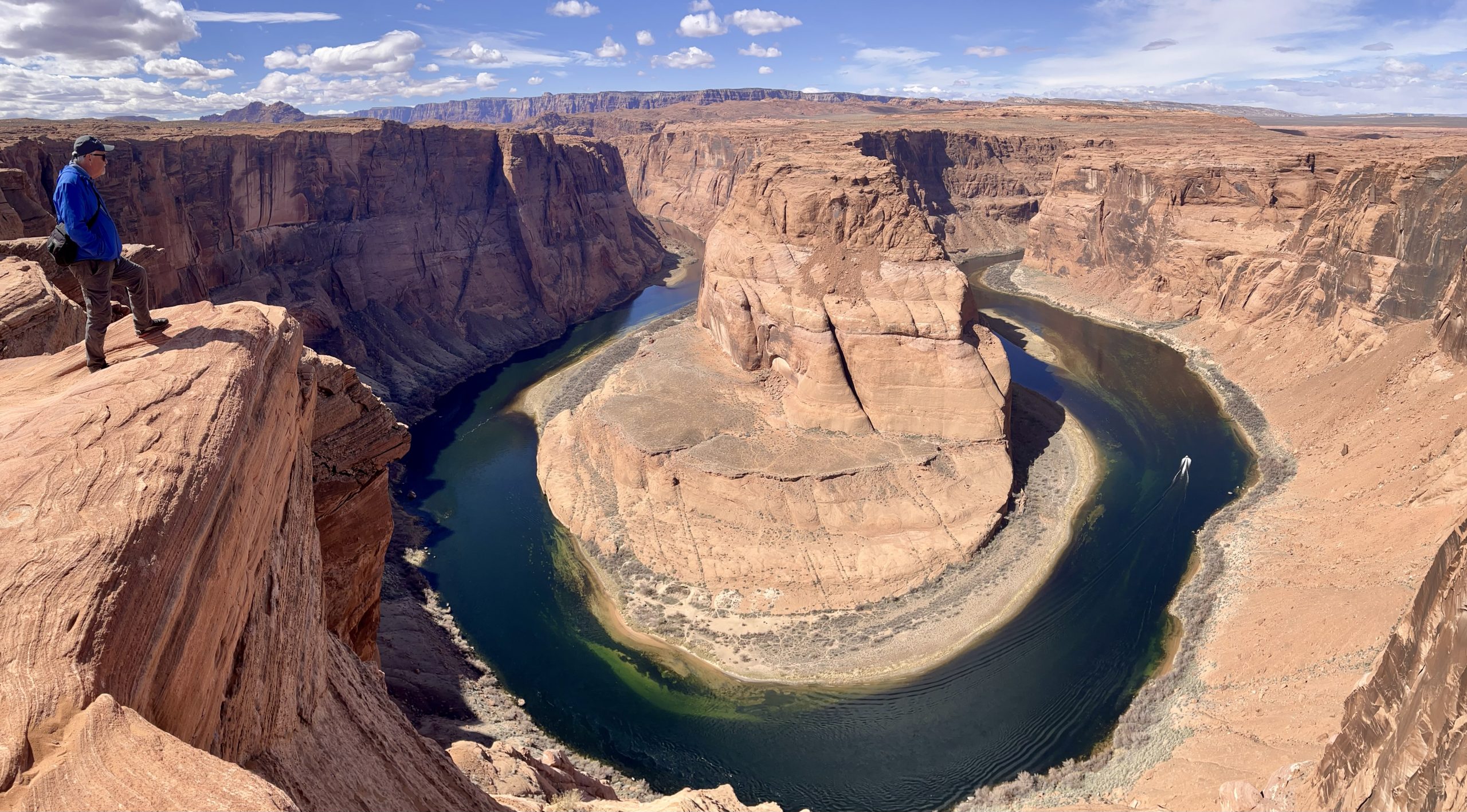 Steep canyon with a river in a U-shape at the base; a man in a blue jacket is standing on top of the cliff outlook.