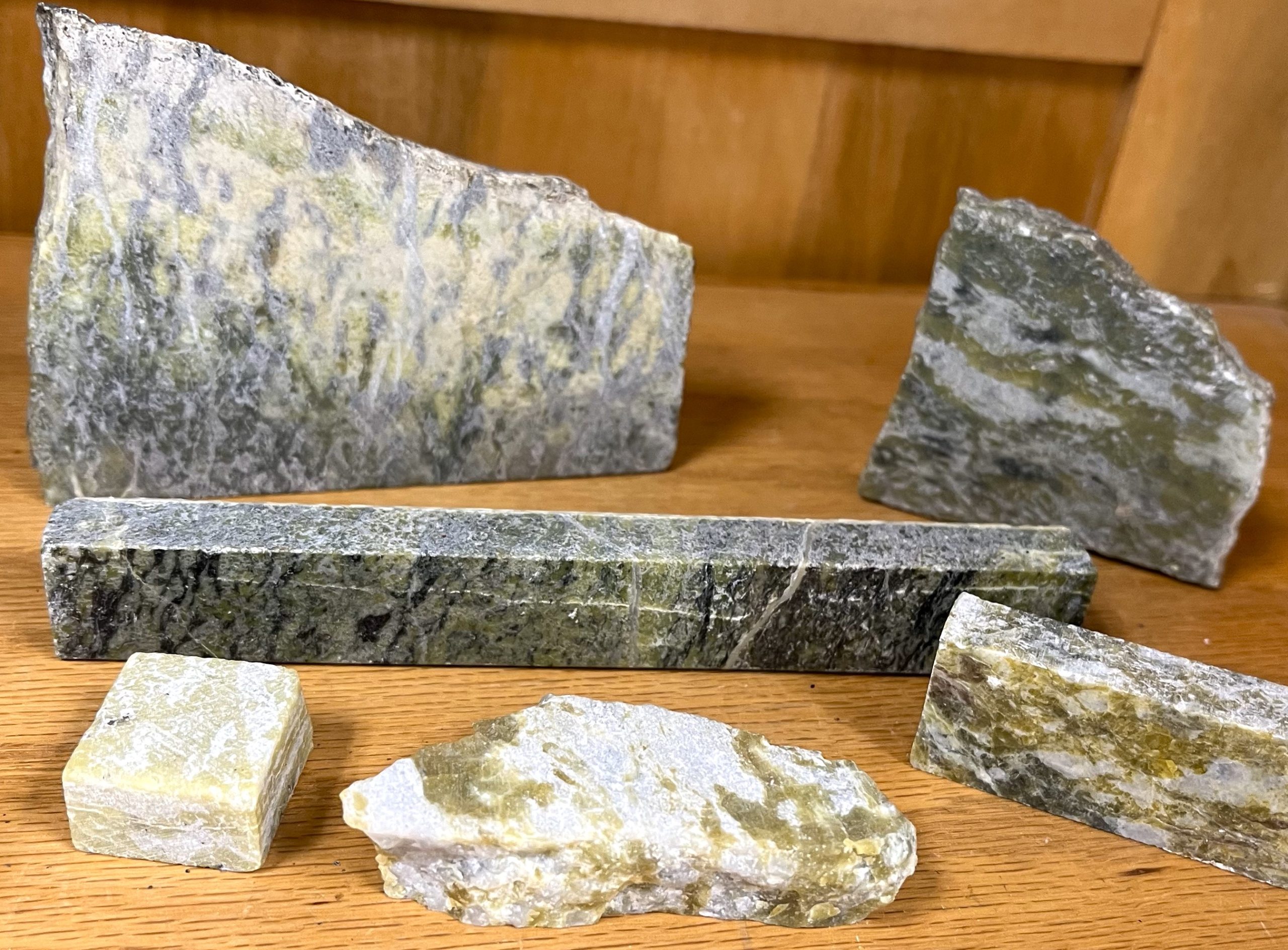 Six pieces of rock on a wooden shelf; all pieces contain shades of green with some being interfingered with white and black.