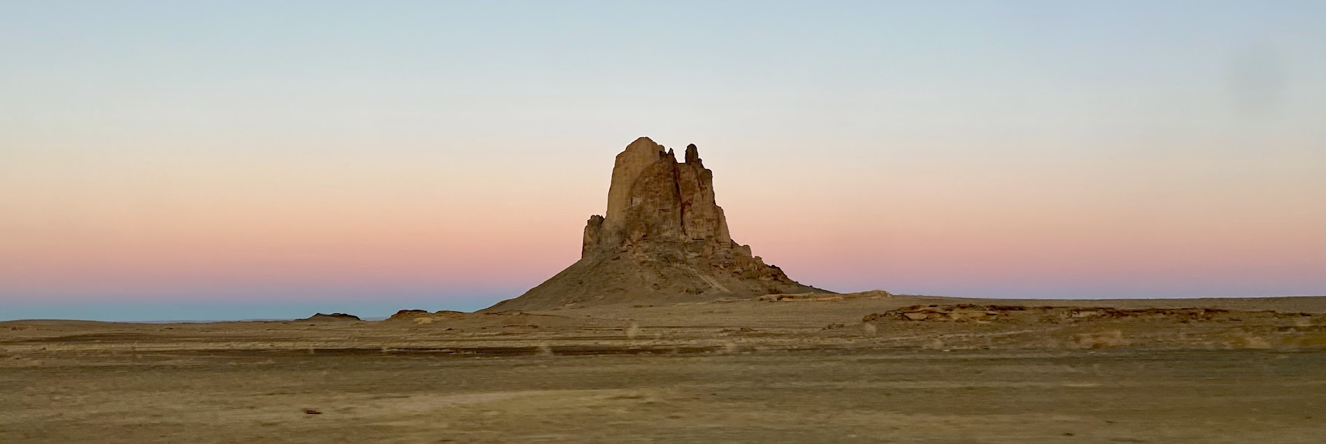 Tan, flat landscape that has a vertical tan-brown rocky outcrop rising high above the surrounding landscape.