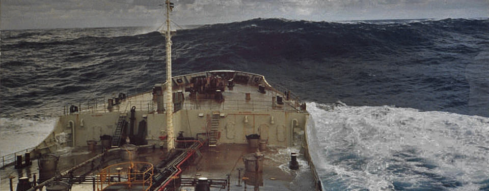 Forward view on top of a ship that's in dark blue water and there is a high steep wave in front of the ship.