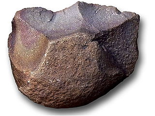 A brown smooth rock that has chips flaked off of it on one side, sharpening it.