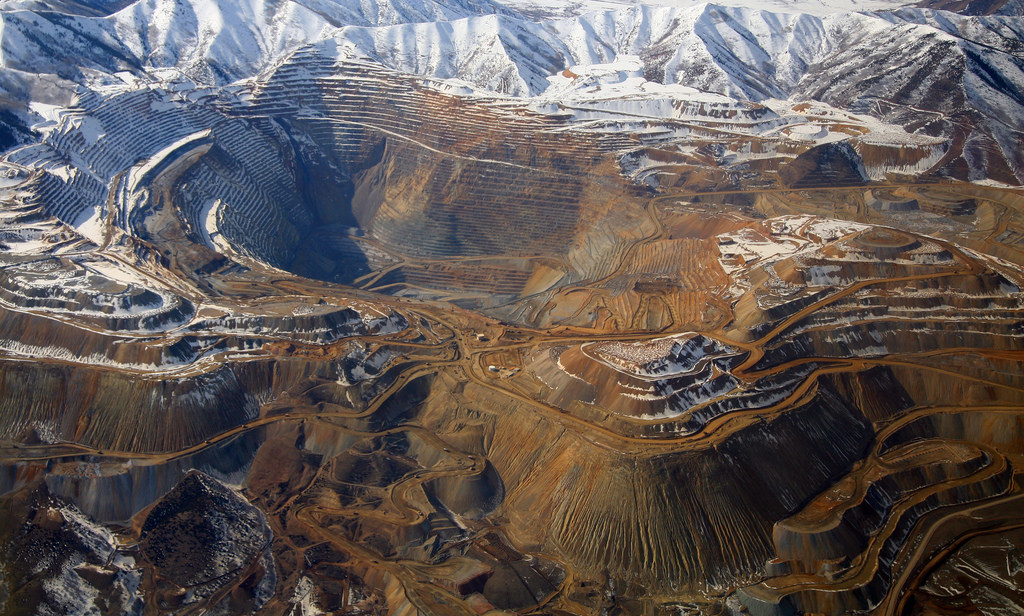 Immense terraced dirt-lined open pit with snow-covered mountains visible around the back of the mine.