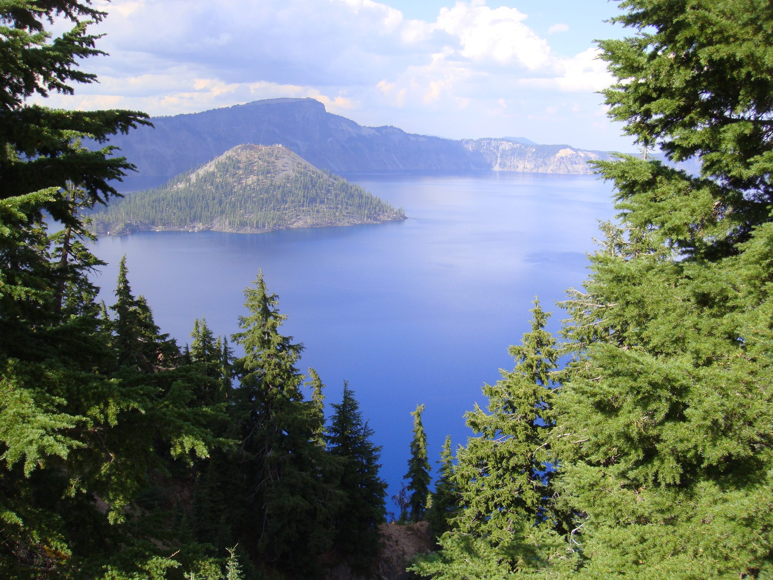 View through a clearing in green trees of a lake with a cone-shaped island in it.