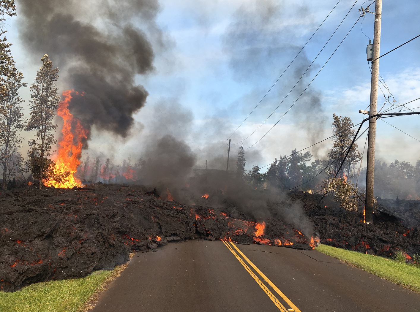 Blocky lava flowing over a road in Hawaii. A tree is on fire on the left-hand side of the photo.