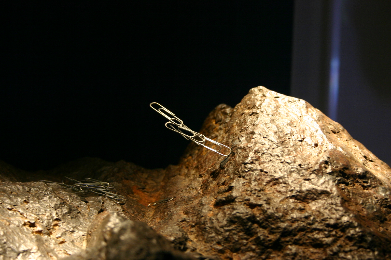 Series of three paper clips together that stick into the air from one end touching the tan sample of lodestone.