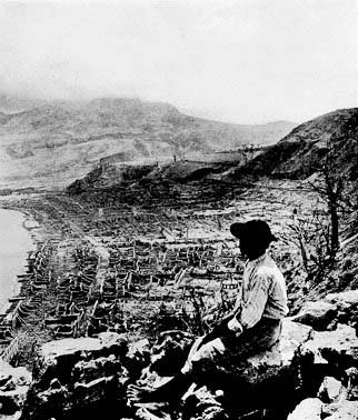 Black and white photo of a man sitting on a ridge, overlooking a destroyed city.