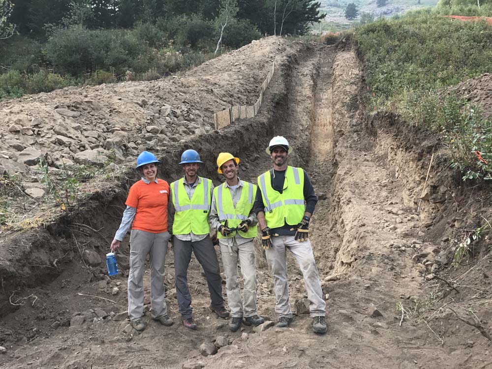 4 people in bright vests stand at the bottom of a trench.