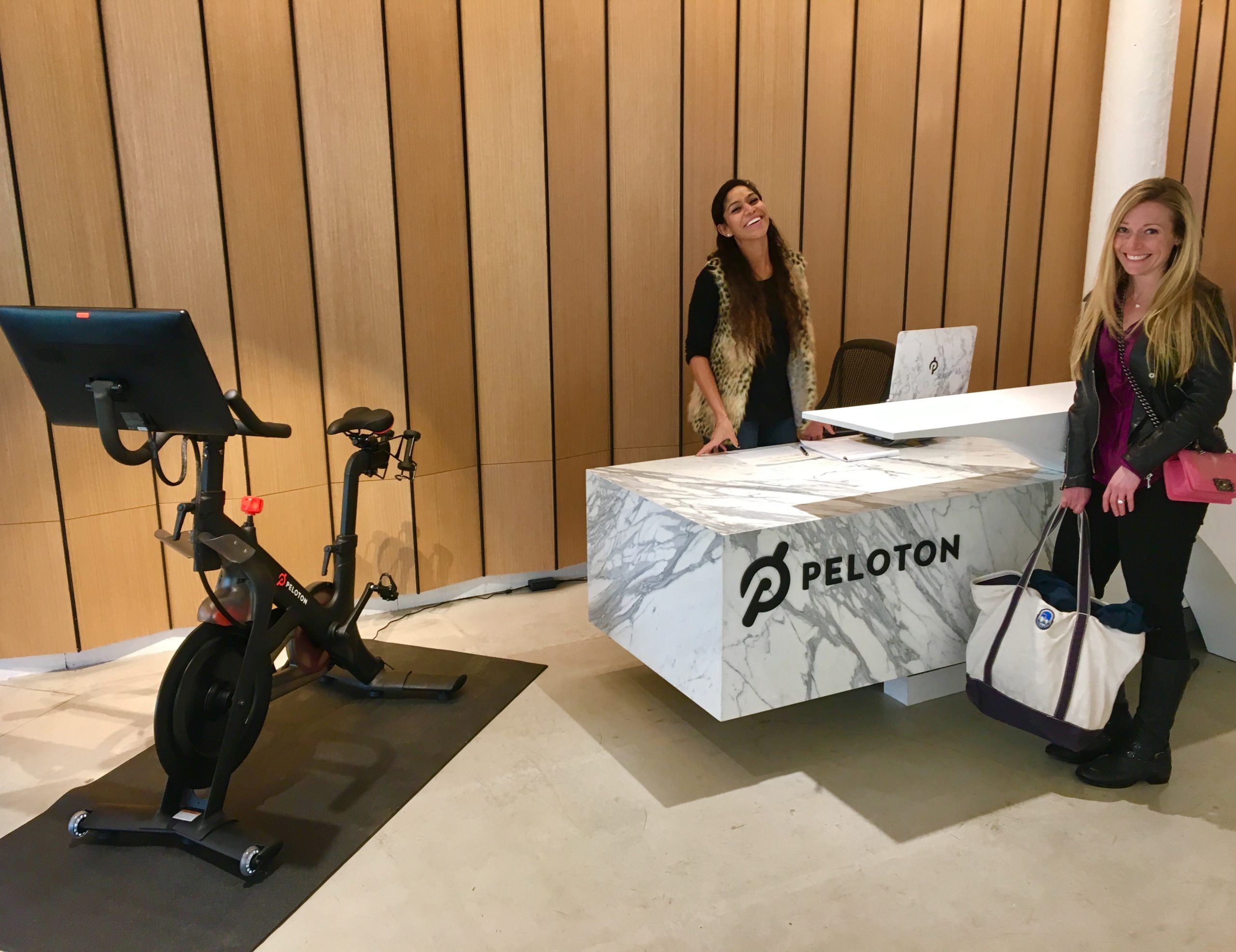 Two women standing near the front desk of a Peloton. There is a Peloton bike beside them