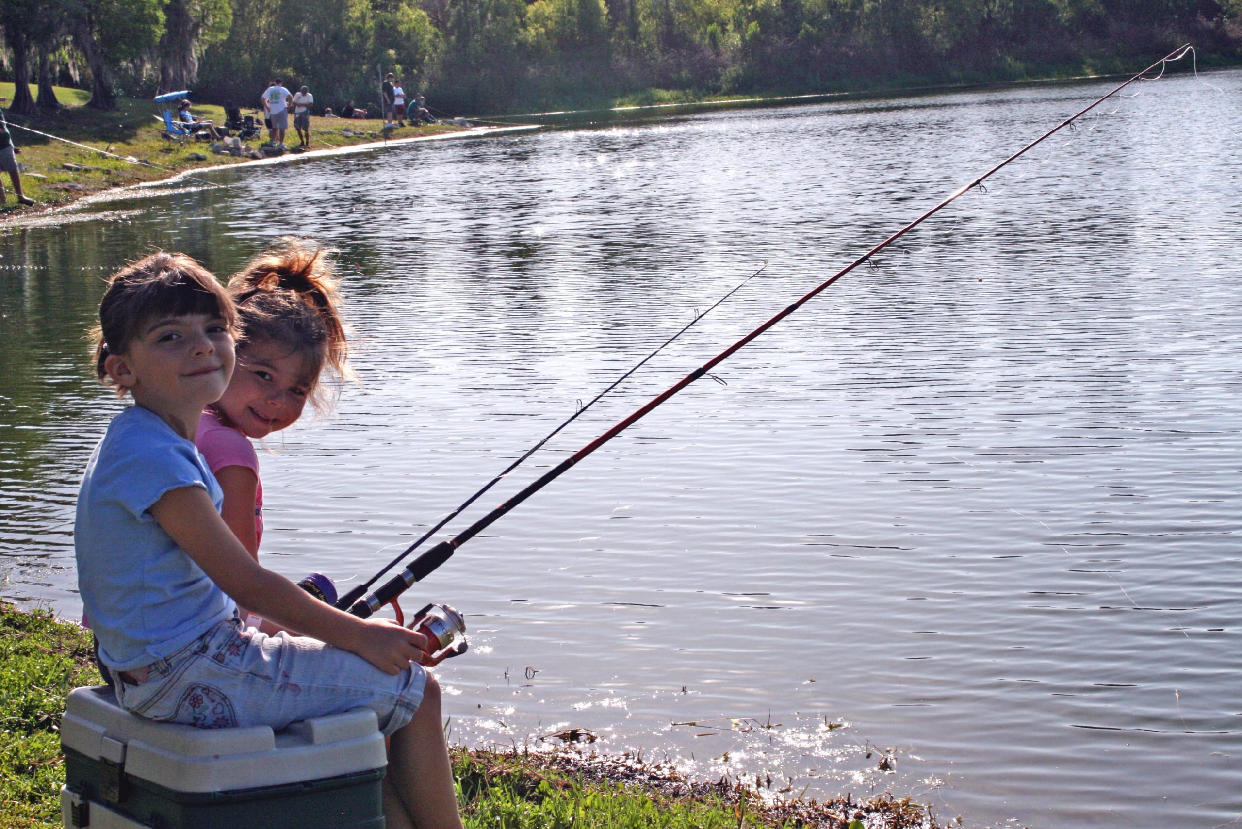 Two children sit on a cooler by a lake, fishing for fun