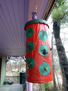 A red cylindrical container hangs from the ceiling of a porch. Green plastic circular openings are spaced evenly along the walls of the cylinder.