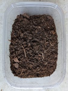 Rectangular plastic container filled halfway with brown organic material of various sizes. Small pieces of bark, sticks, and fine dirt are visible.