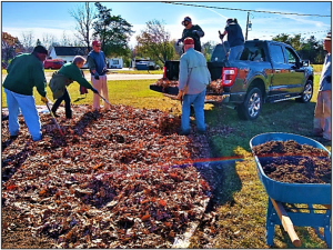 Group of people stands around a large area of dirt and leaves amid a green lawn.