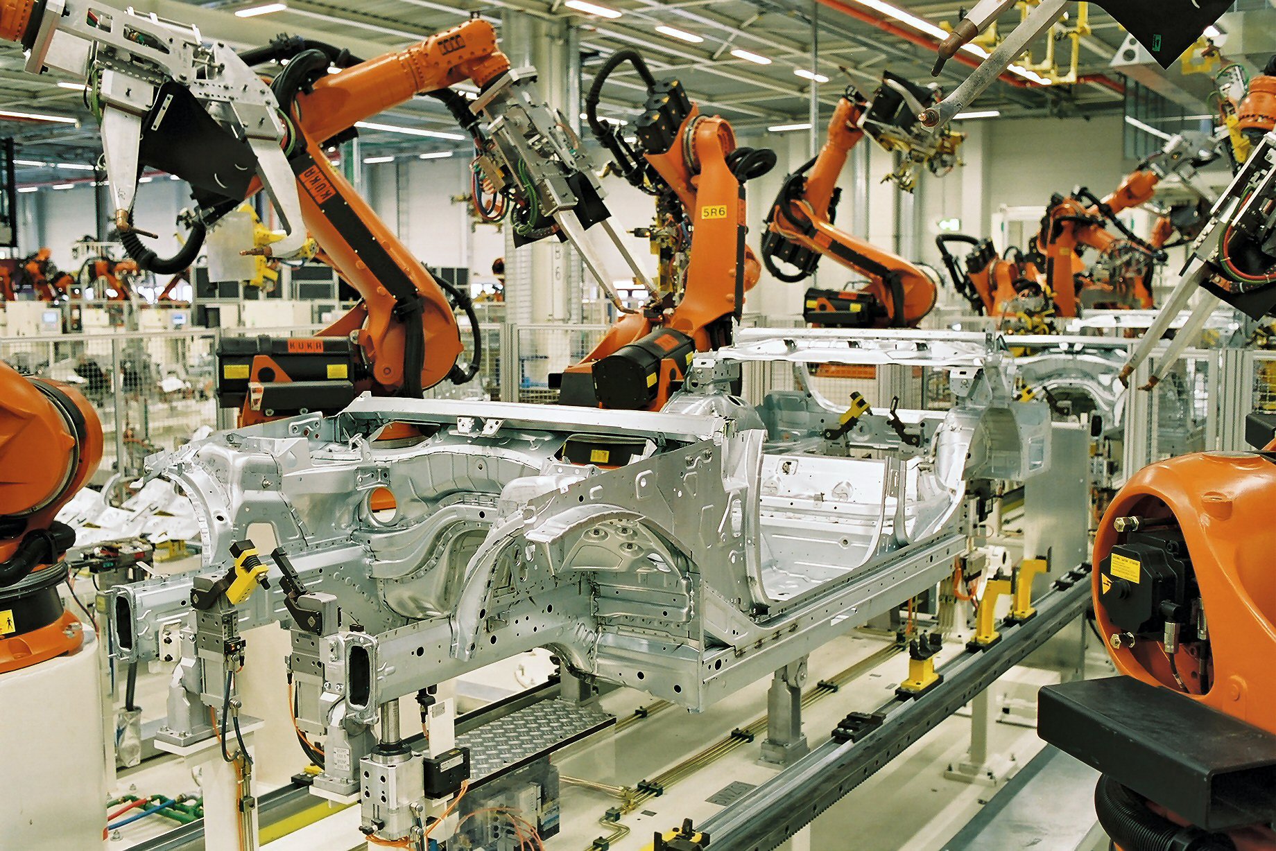 A photograph of seven orange mechanical arms in a warehouse, working on the beginnings of automobile bodies in an assembly line.