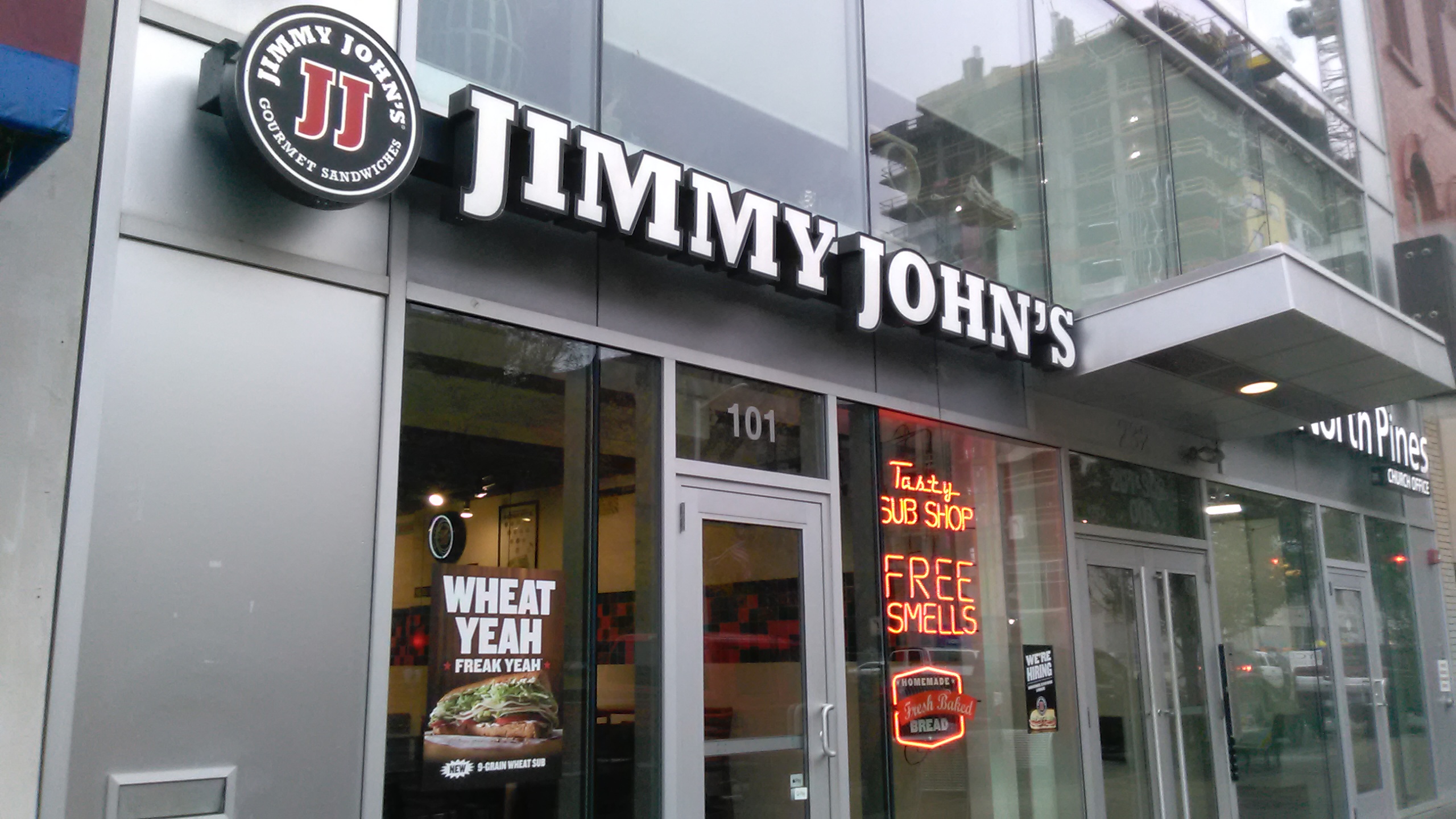 Outside view of a modern Jimmy Johns with neon signs reading "tasy sub shop" and "free smells"