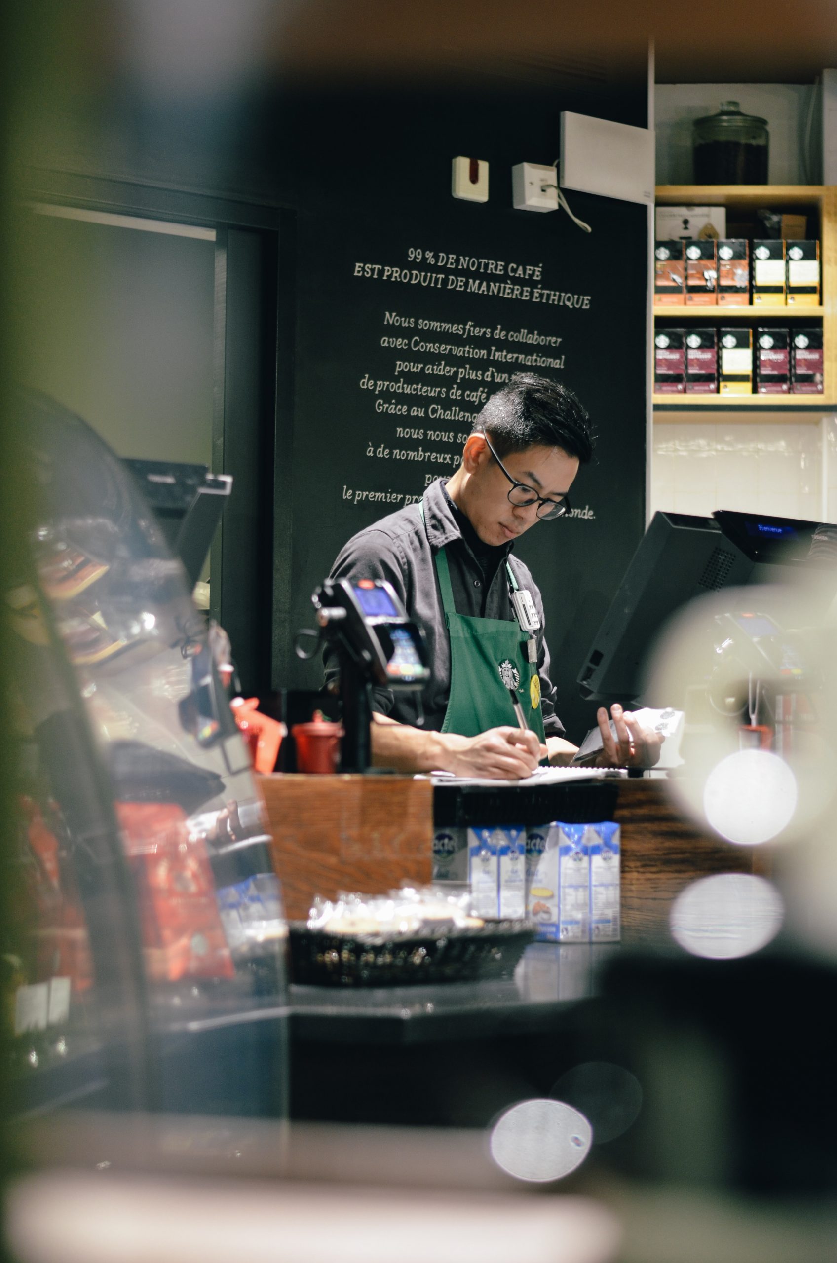 A male Starbucks barista stands behind the counter writing on paper. He is wearing a green Starbucks apron and is standing behind the cash register.