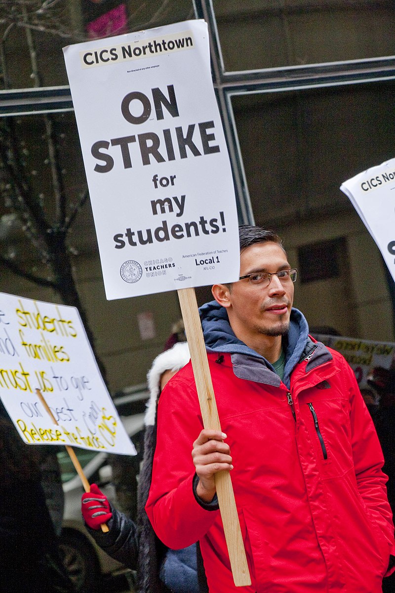 A photograph of a teacher protesting in a crowd holding a large, square sign reading “On strike for my students!”