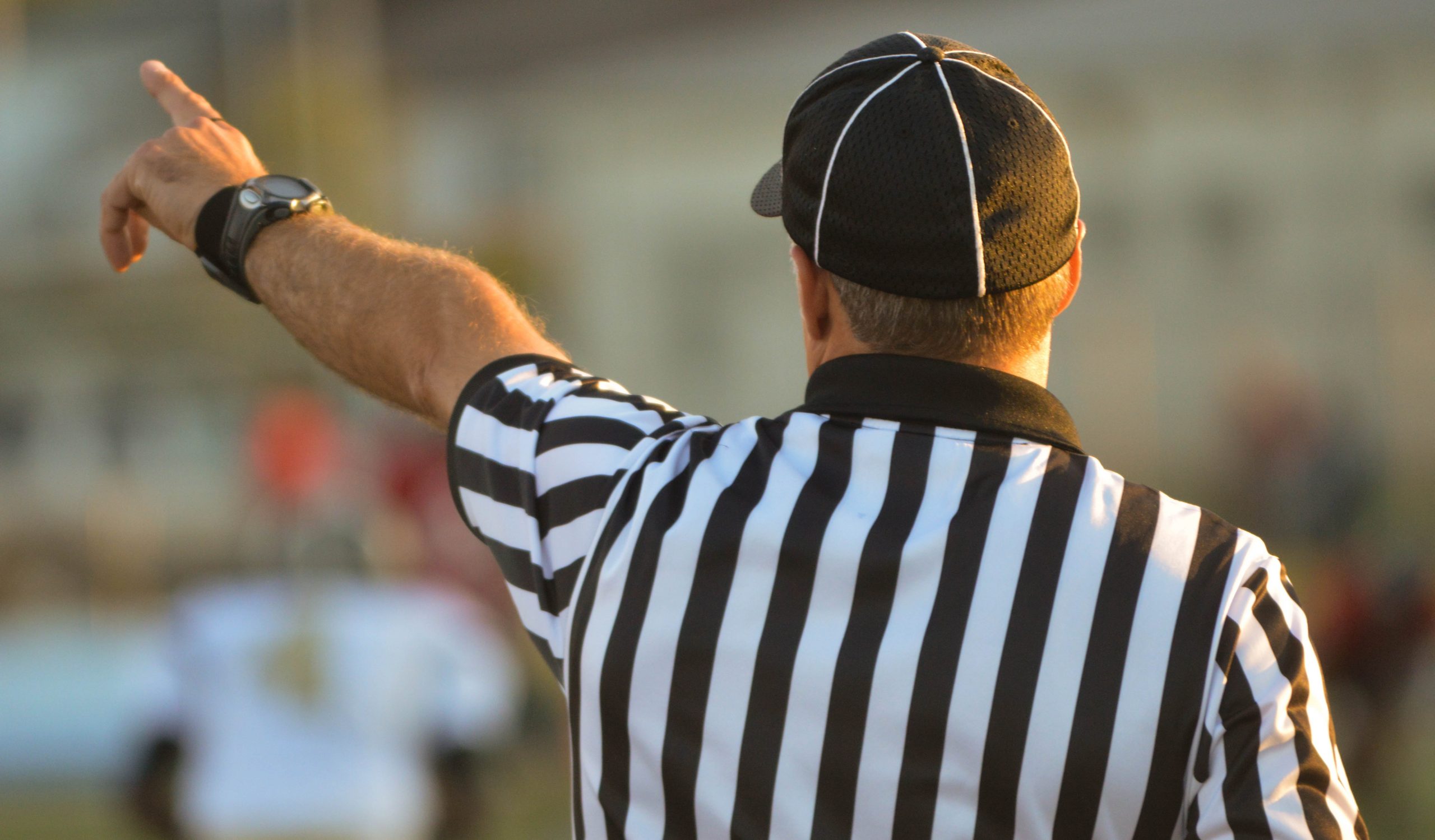 Photograph of a referee with his back facing the camera. He's wearing a black and white striped shirt, a black hat, and a watch. He's pointing to something off camera.