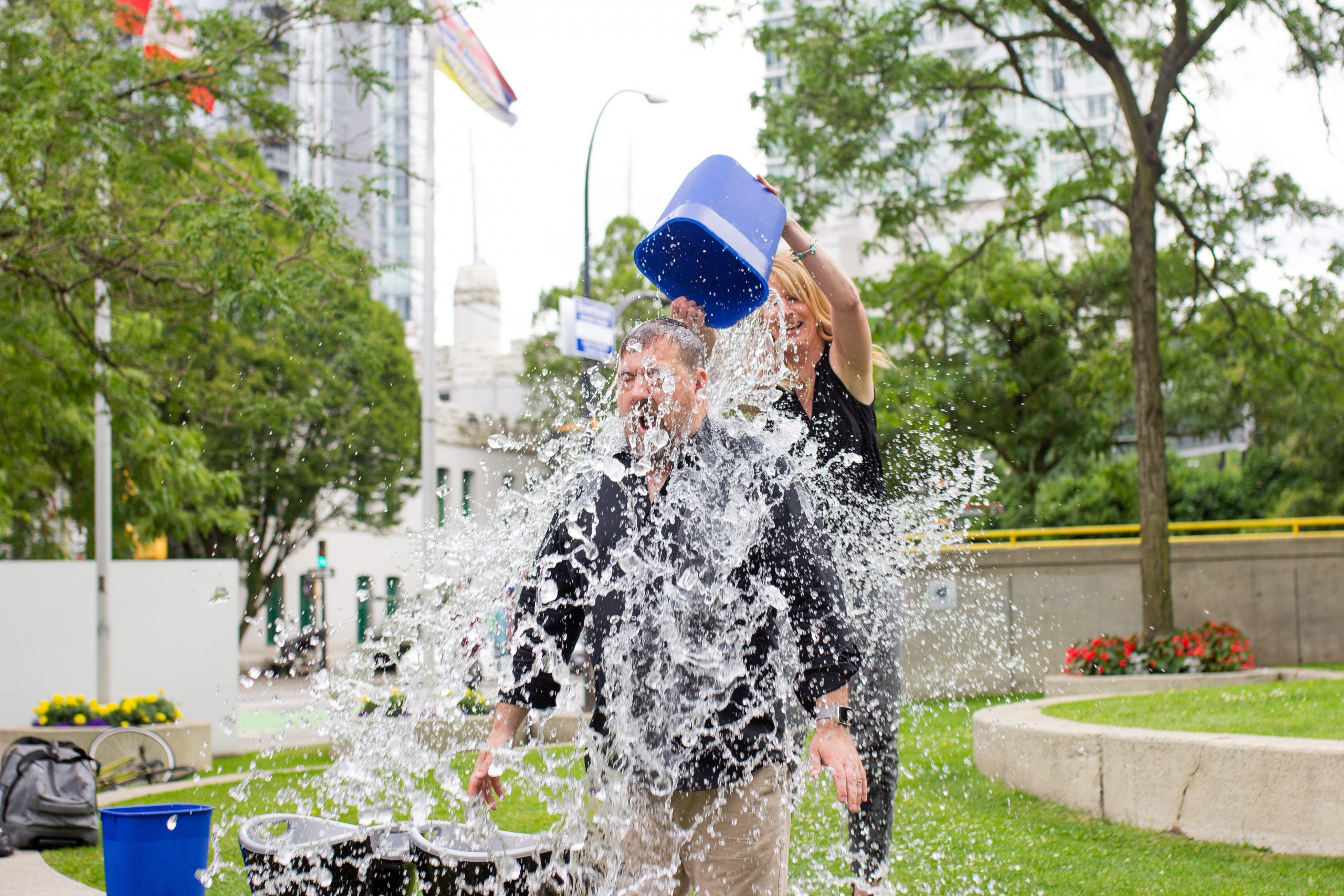 Photo of a woman pouring a bucket of water over a man's head outside in a park.