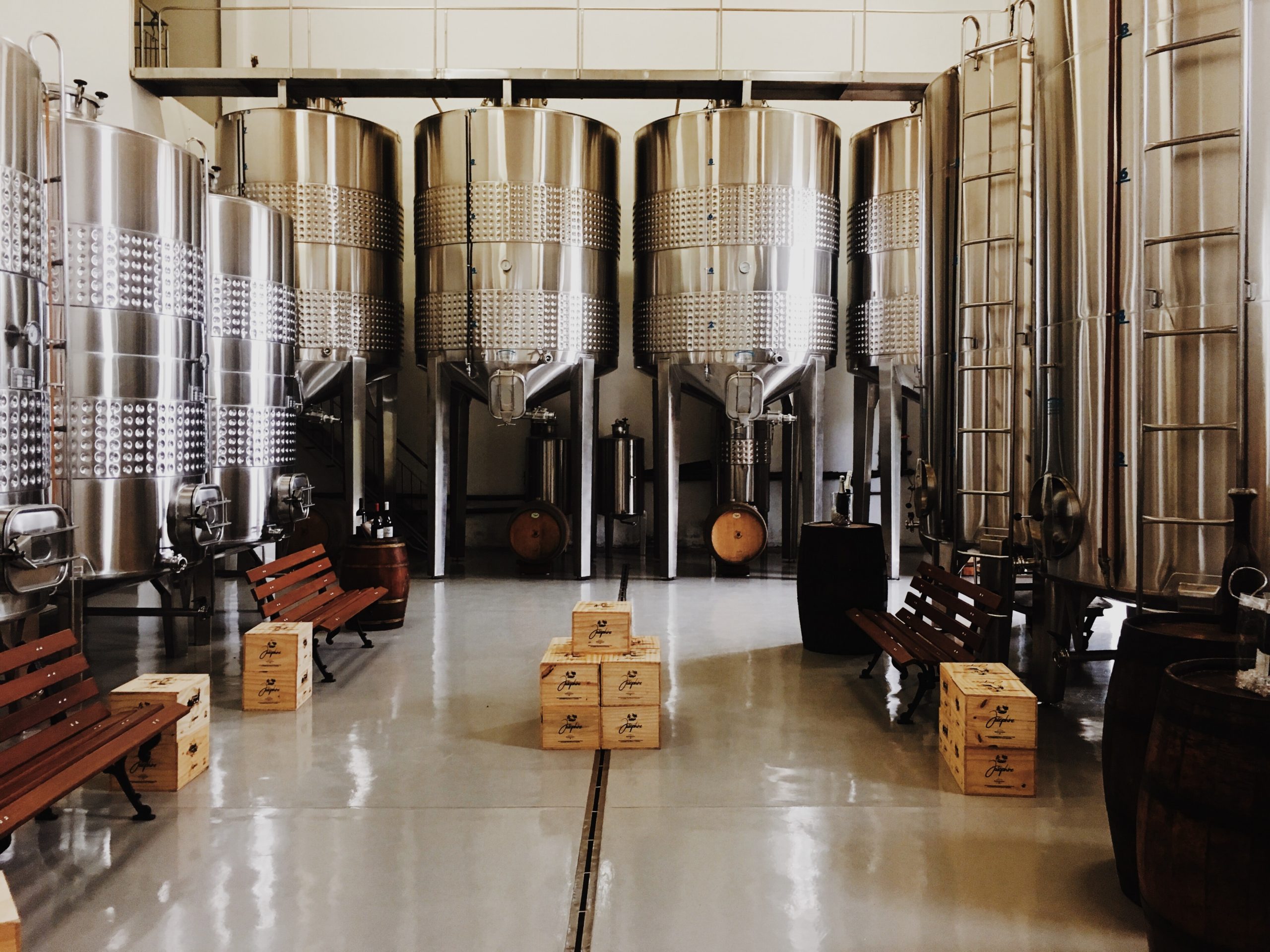 9 large, shiny, steel cylindical containers stand upright in a room with benches, boxes, and barrels on the floor.